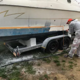 Pracownik w kombinezonie ochronnym szlifuje kadłub łodzi umieszczonej na przyczepie, przygotowując ją do malowania.