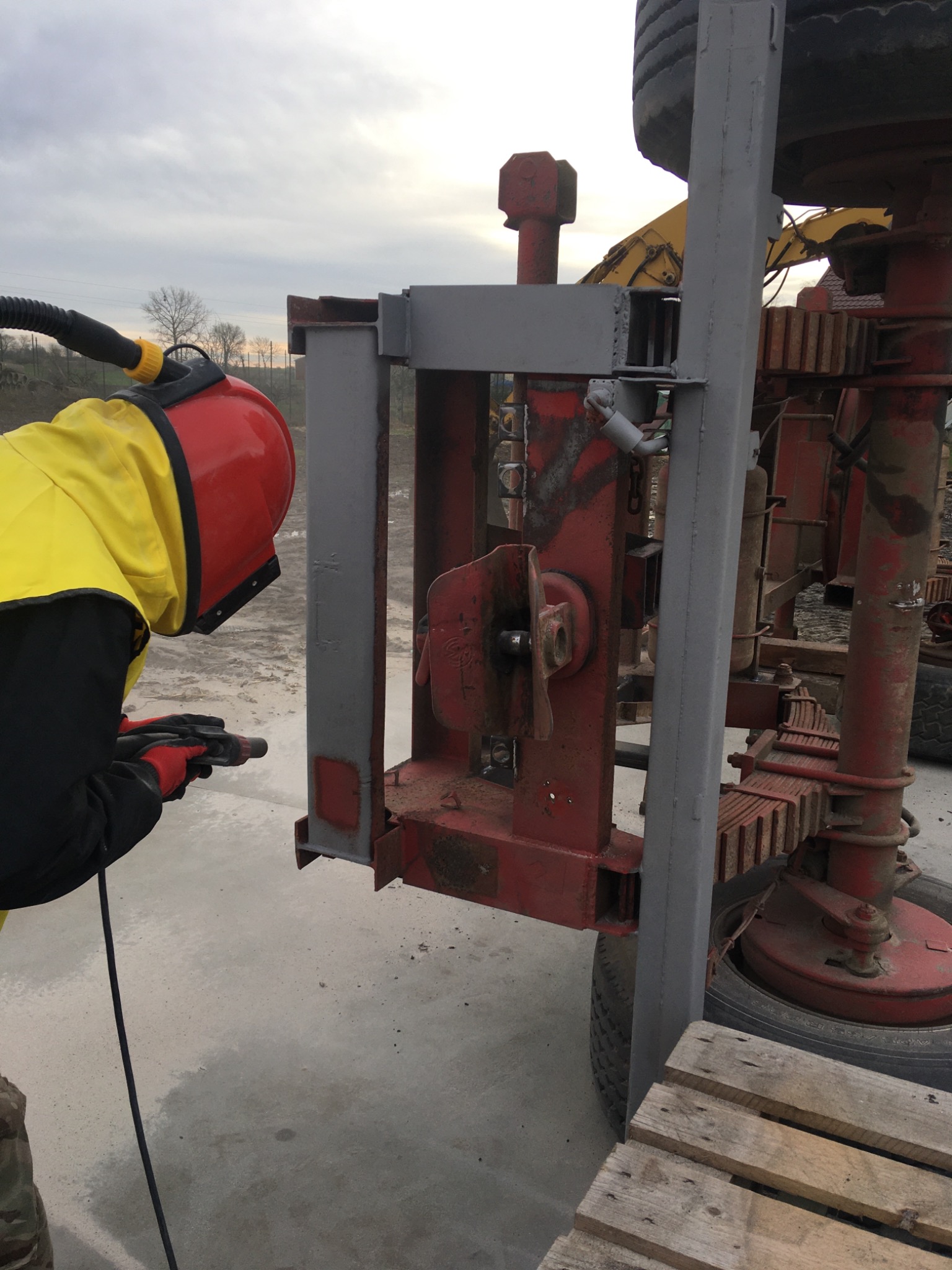 Pracownik w kombinezonie ochronnym i hełmie z maską wykonuje czyszczenie strumieniowo-ścierne metalowej konstrukcji na zewnątrz, z widoczną maszyną w tle.