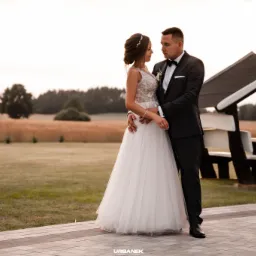 Para młoda w objęciach na tle pola, w tle altanka z białymi ławkami, panna młoda w sukni z koronkową górą i tiulowym dołem, pan młody w czarnym garniturze i muszce.