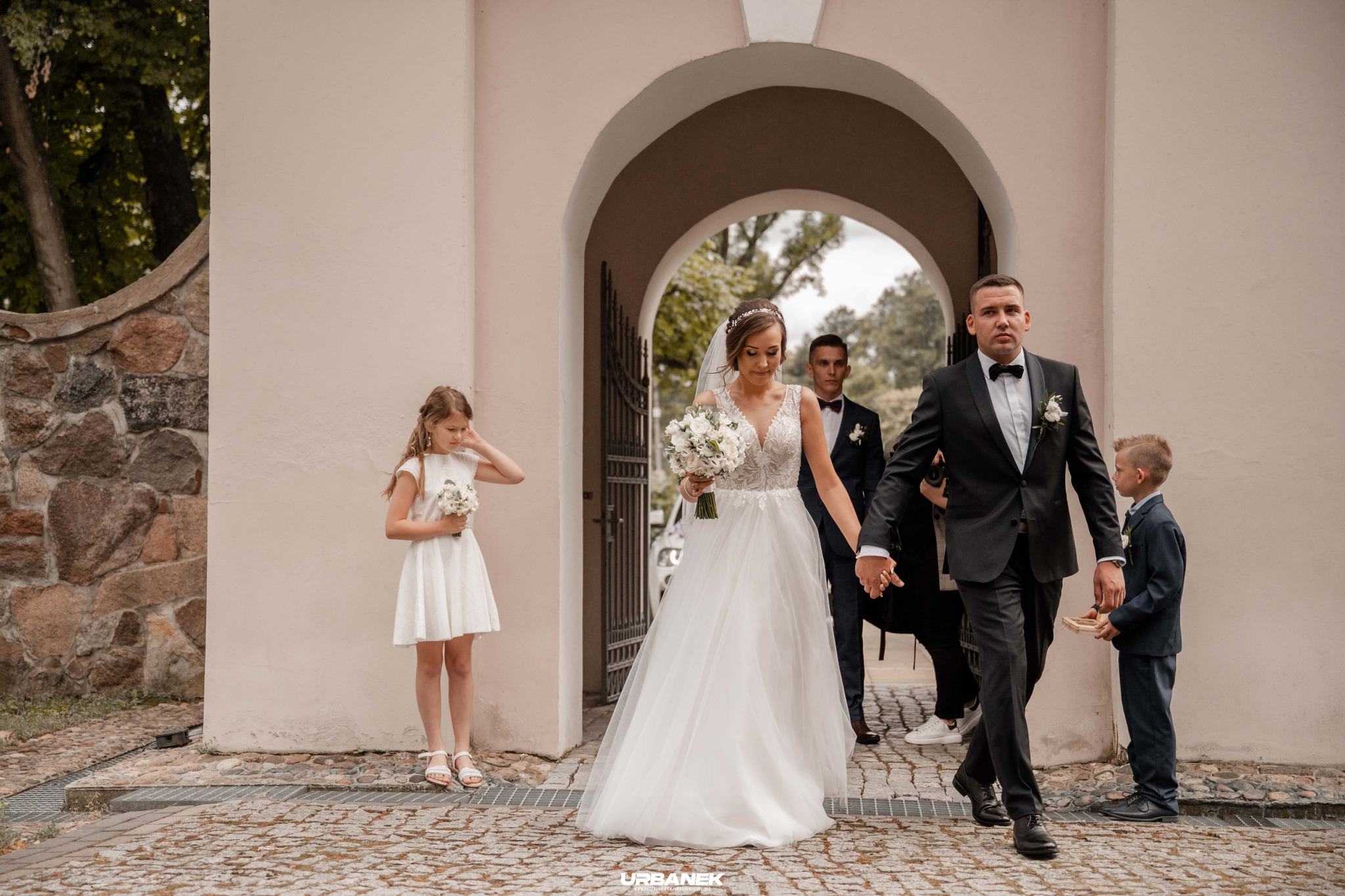 Para młoda wychodzi z kościoła, trzymając się za ręce, w tle drużba i chłopiec niosący obrączki, obok dziewczynka w białej sukience.