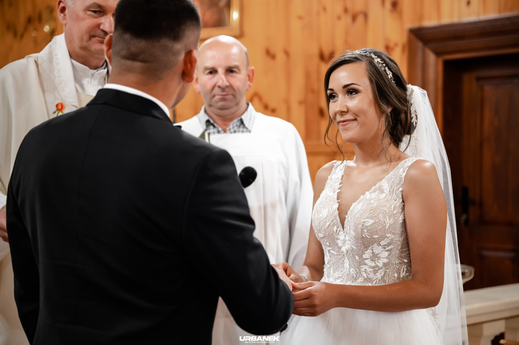 Panna młoda w białej sukni z koronkowymi aplikacjami i welonem, uśmiechnięta podczas ceremonii ślubnej, trzymająca dłoń pana młodego w czarnym garniturze na tle drewnianej ściany kościoła, widoczni...