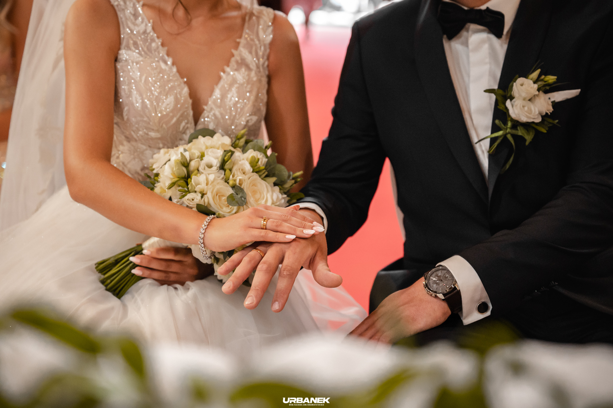 Dłonie pary młodej z obrączkami spoczywają na sobie, panna młoda trzyma bukiet białych róż i frezji, a pan młody ma na nadgarstku elegancki zegarek.