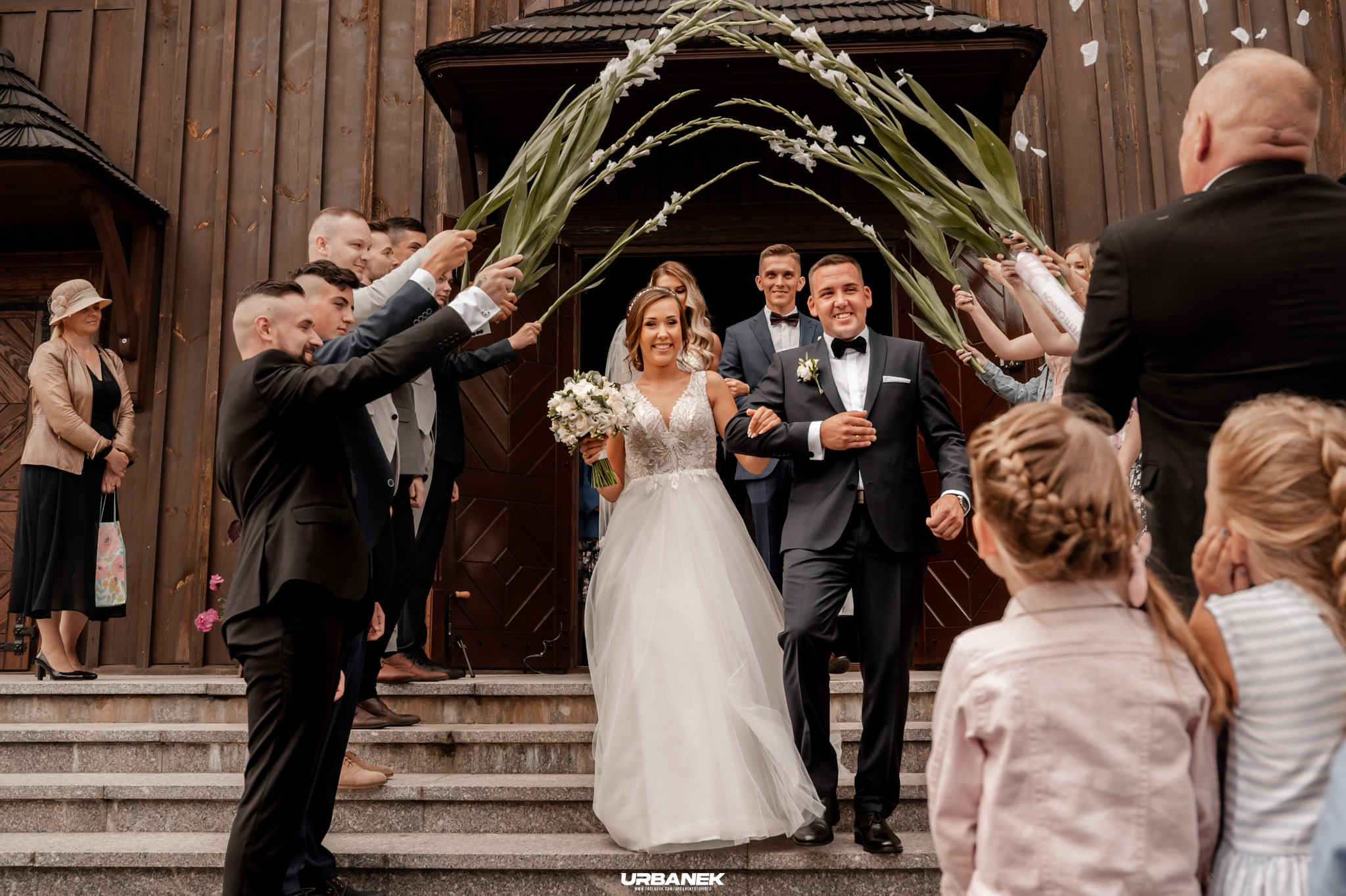 Para młoda wychodzi z drewnianego kościoła pod szpalerem z zielonych gałęzi i białych kwiatów, obsypywana konfetti, na schodach czekają dzieci.