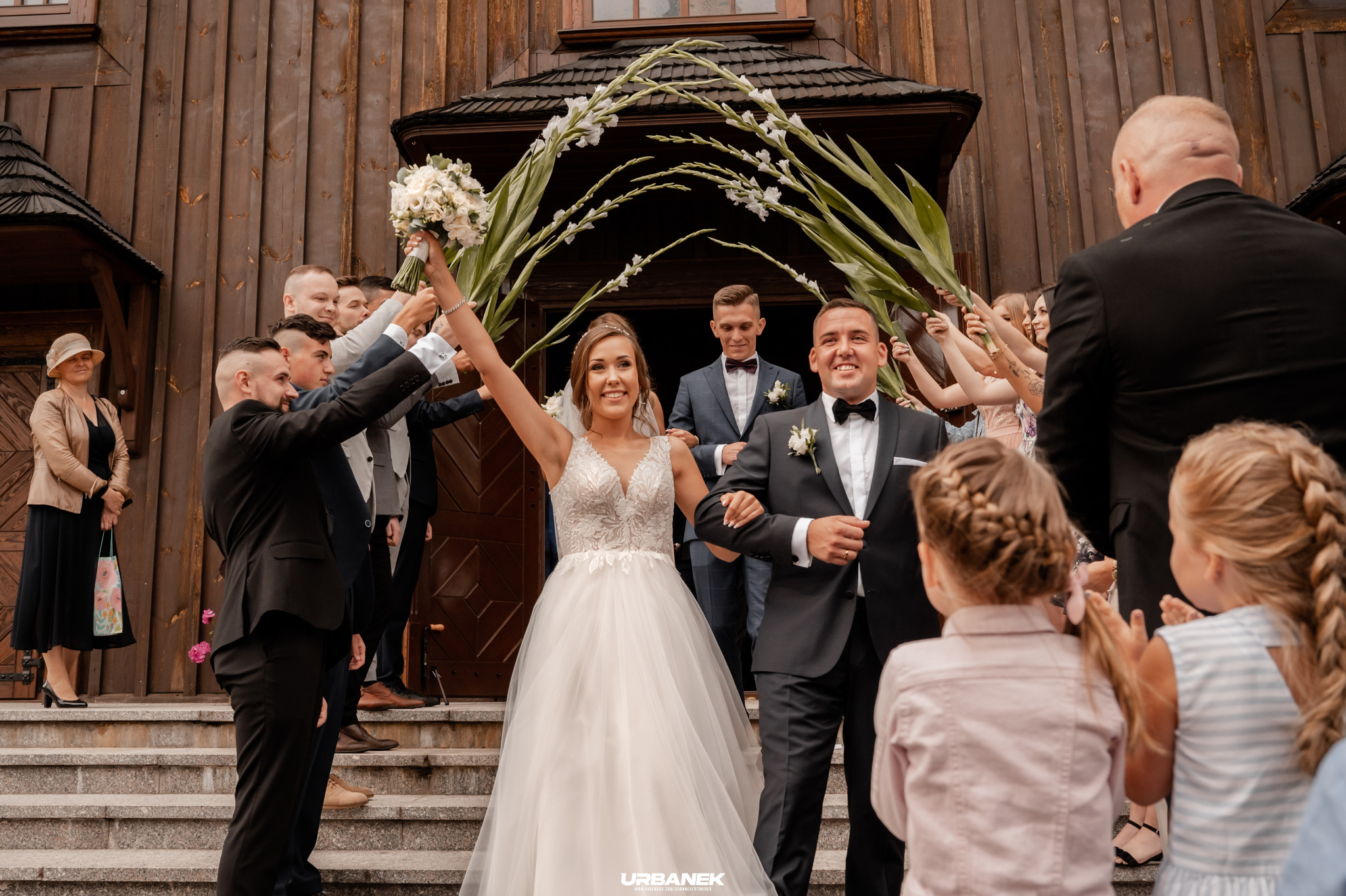Para młoda wychodzi z drewnianego kościoła pod szpalerem z kwiatów i zieleni, otoczona gośćmi weselnymi.