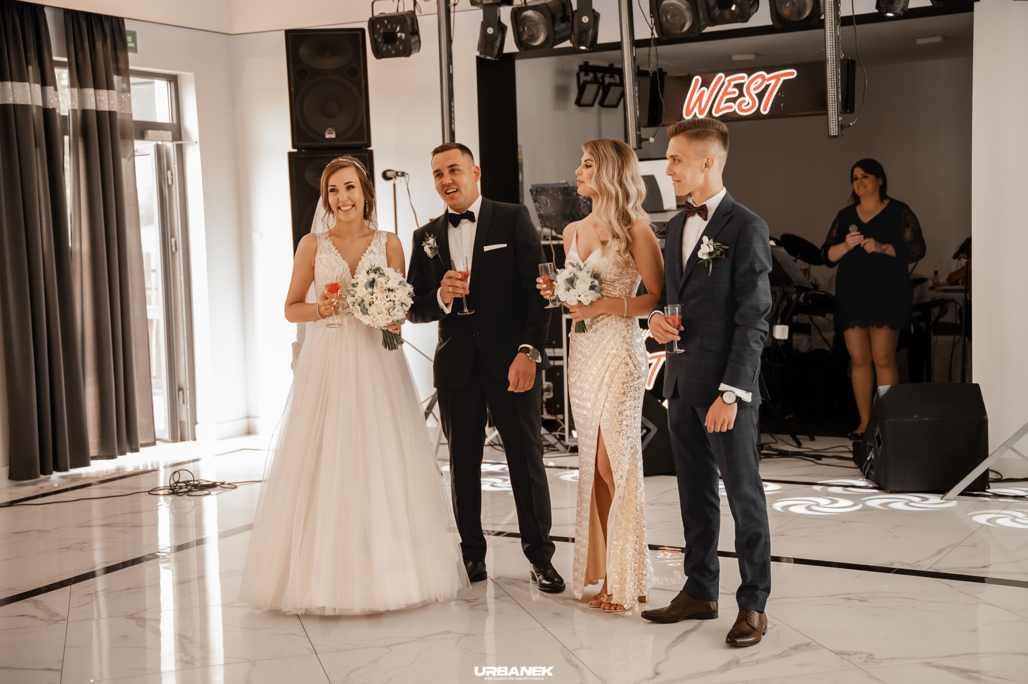 Para młoda i goście w eleganckich strojach wznoszą toast w sali weselnej z neonowym napisem 'WEST' w tle.