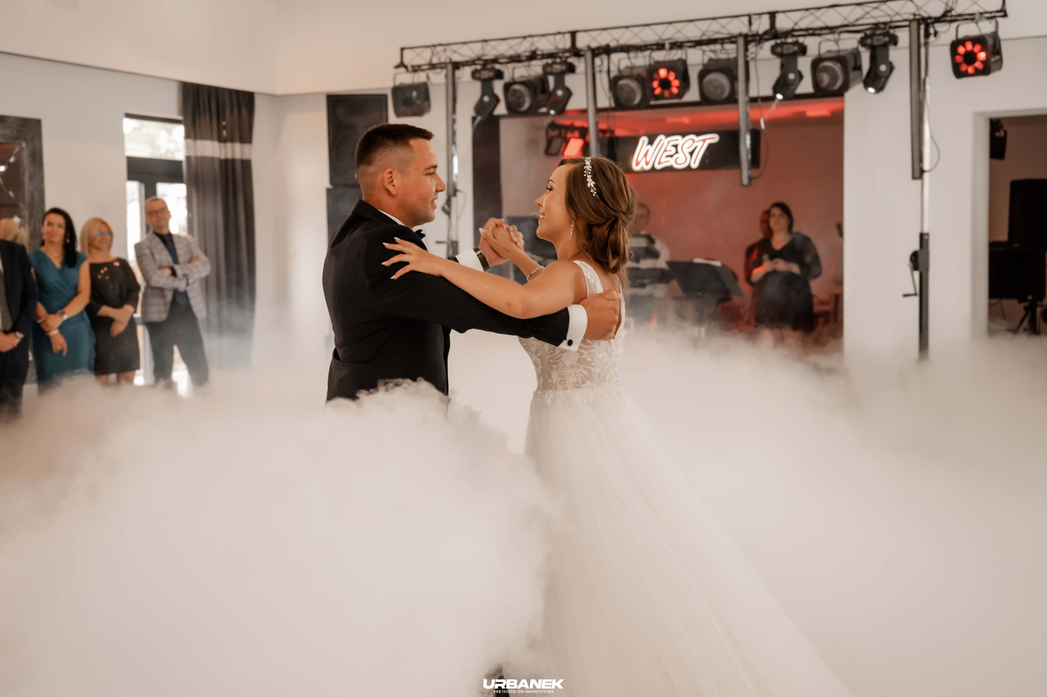 Para młoda w tańcu podczas wesela, otoczona sztuczną mgłą na parkiecie, w tle goście i oświetlenie sceniczne.