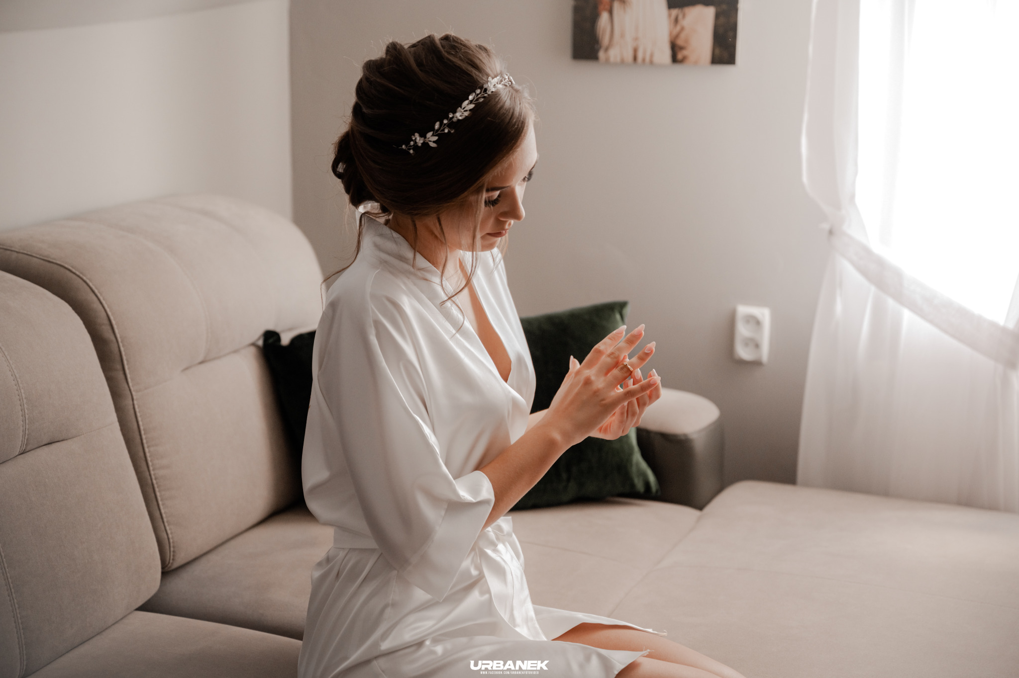 Panna młoda w białym szlafroku, z delikatną ozdobą we włosach, siedzi na beżowej kanapie i ogląda pierścionek zaręczynowy na palcu, przygotowując się do ślubu.