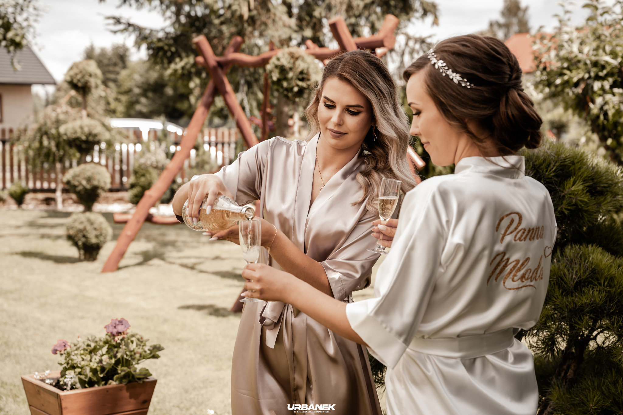 Dwie kobiety w szlafrokach w ogrodzie, jedna nalewa szampana do kieliszków, na plecach szlafroka drugiej kobiety widoczny napis 'Panna Młoda'.