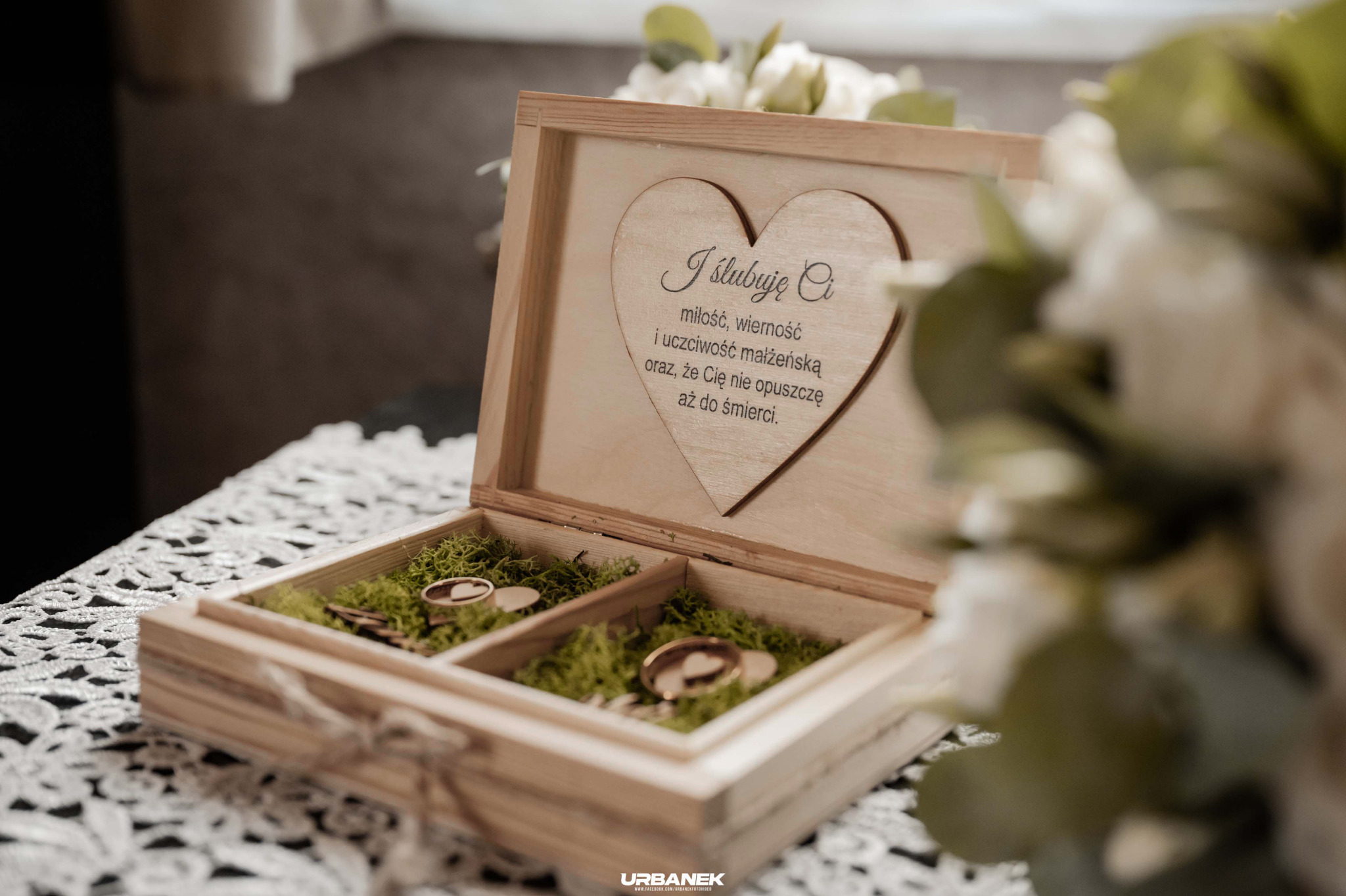 Drewniana szkatułka z wygrawerowanym sercem i tekstem przysięgi małżeńskiej, wewnątrz mech i złote obrączki ślubne, na koronkowej serwetce, w tle bukiet kwiatów.