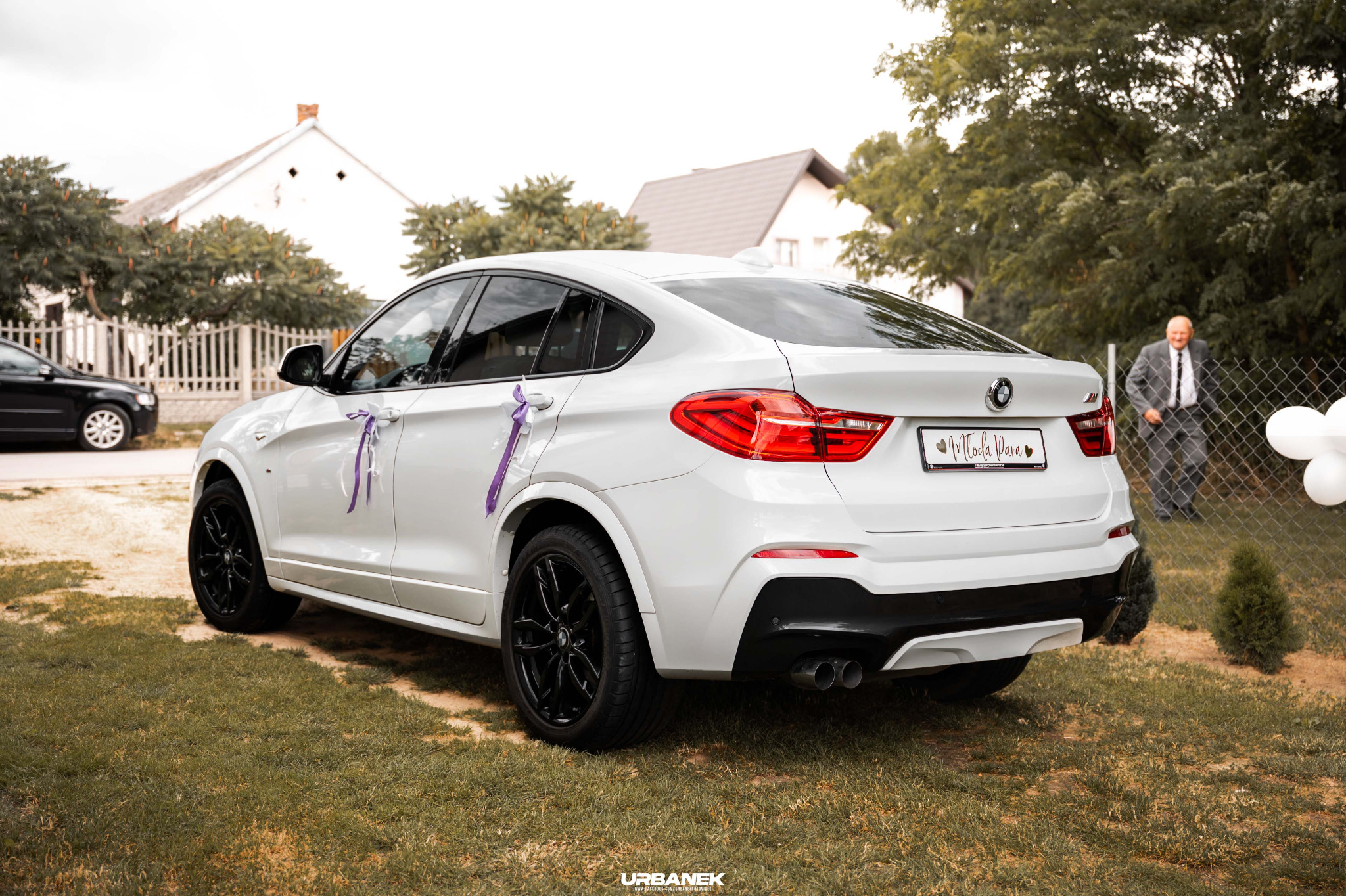 Białe BMW X4 udekorowane fioletowymi wstążkami, tablica rejestracyjna z napisem 'Młoda Para', w tle dom i mężczyzna w garniturze.