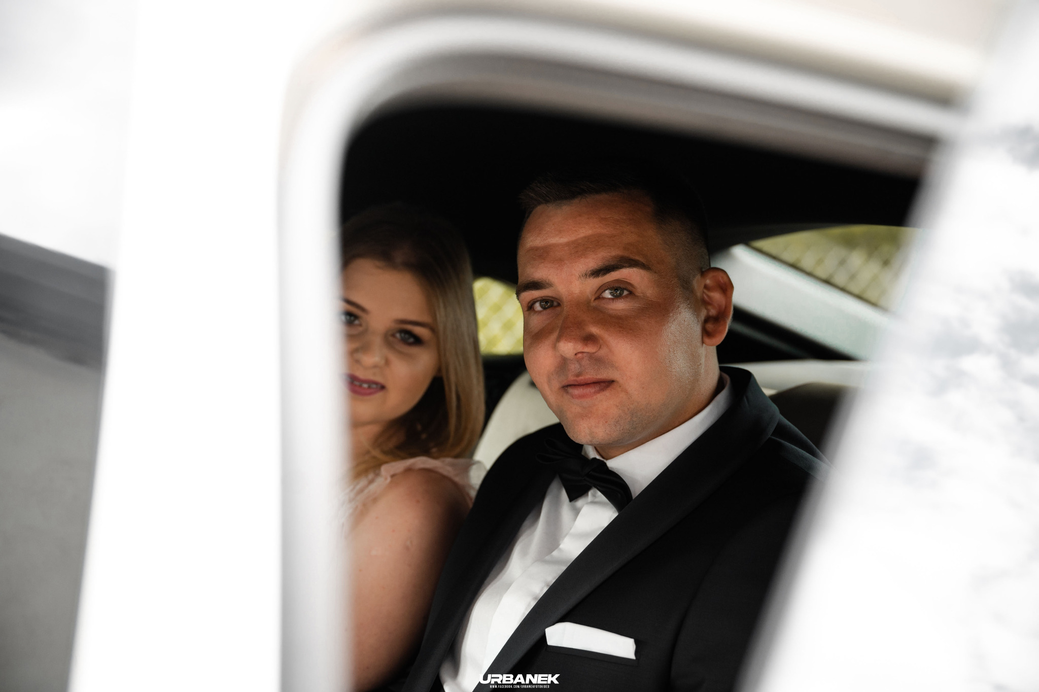 Para młoda w samochodzie: pan młody w czarnym smokingu z muchą, panna młoda z blond włosami w tle, widok przez otwarte drzwi auta.