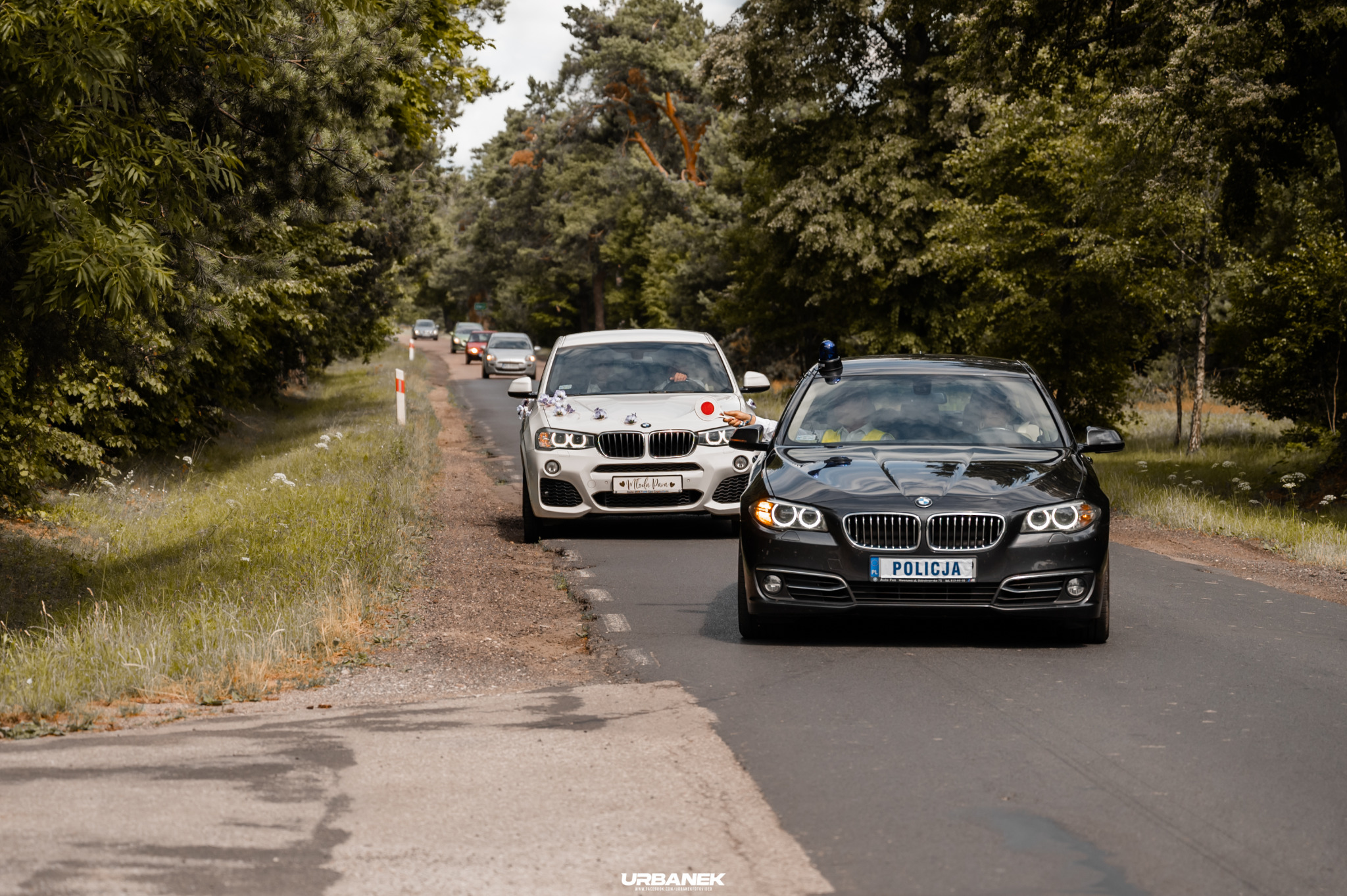 Kolumna samochodów na drodze, w tym czarne BMW z napisem 'Policja' na tablicy rejestracyjnej oraz białe BMW udekorowane kwiatami i tabliczką 'Młoda Para'.