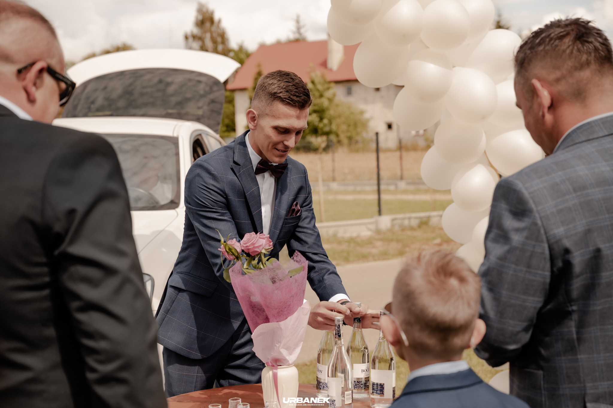 Elegancko ubrany mężczyzna w muszce, z bukietem różowych róż, otwiera butelkę wódki na stole podczas uroczystości na zewnątrz, z białymi balonami w tle i otwartym bagażnikiem białego samochodu.