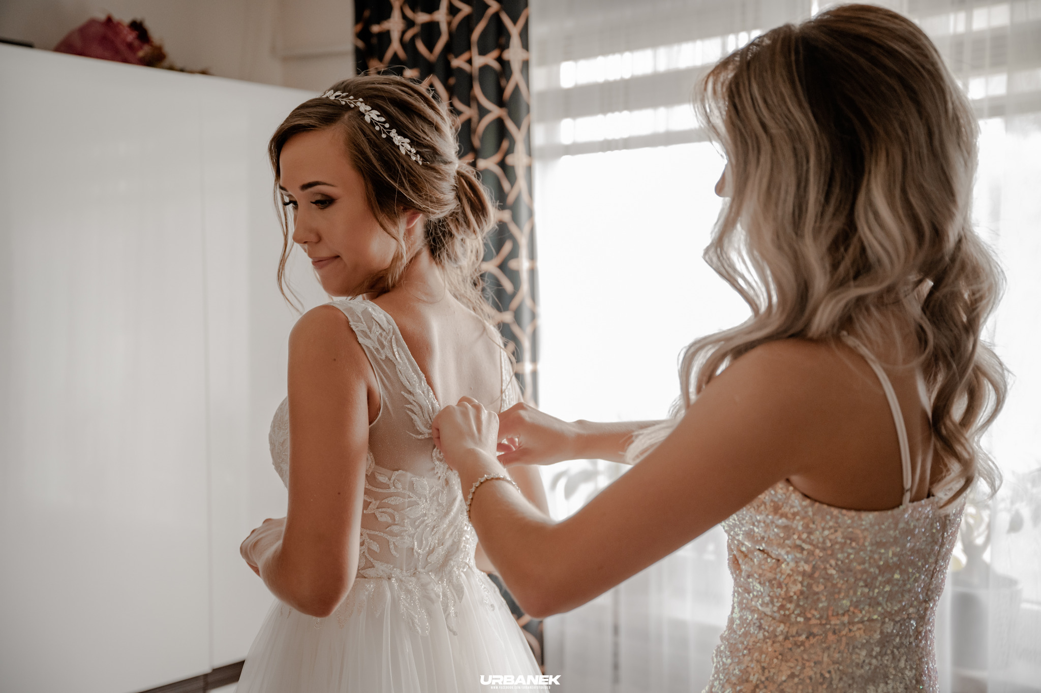 Panna młoda w białej sukni ślubnej z tiulowym dołem, dopinana przez druhnę w cekinowej sukience na tle okna.