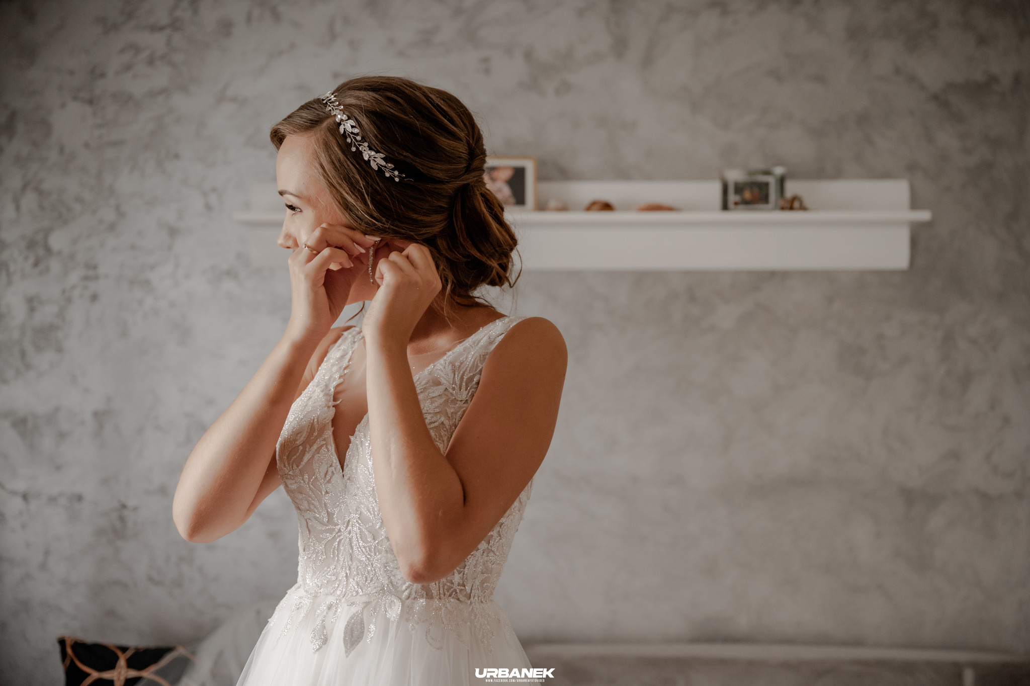 Panna młoda w białej sukni z koronkowymi zdobieniami, z profilu, zapina kolczyk, w tle półka z ramkami ze zdjęciami.