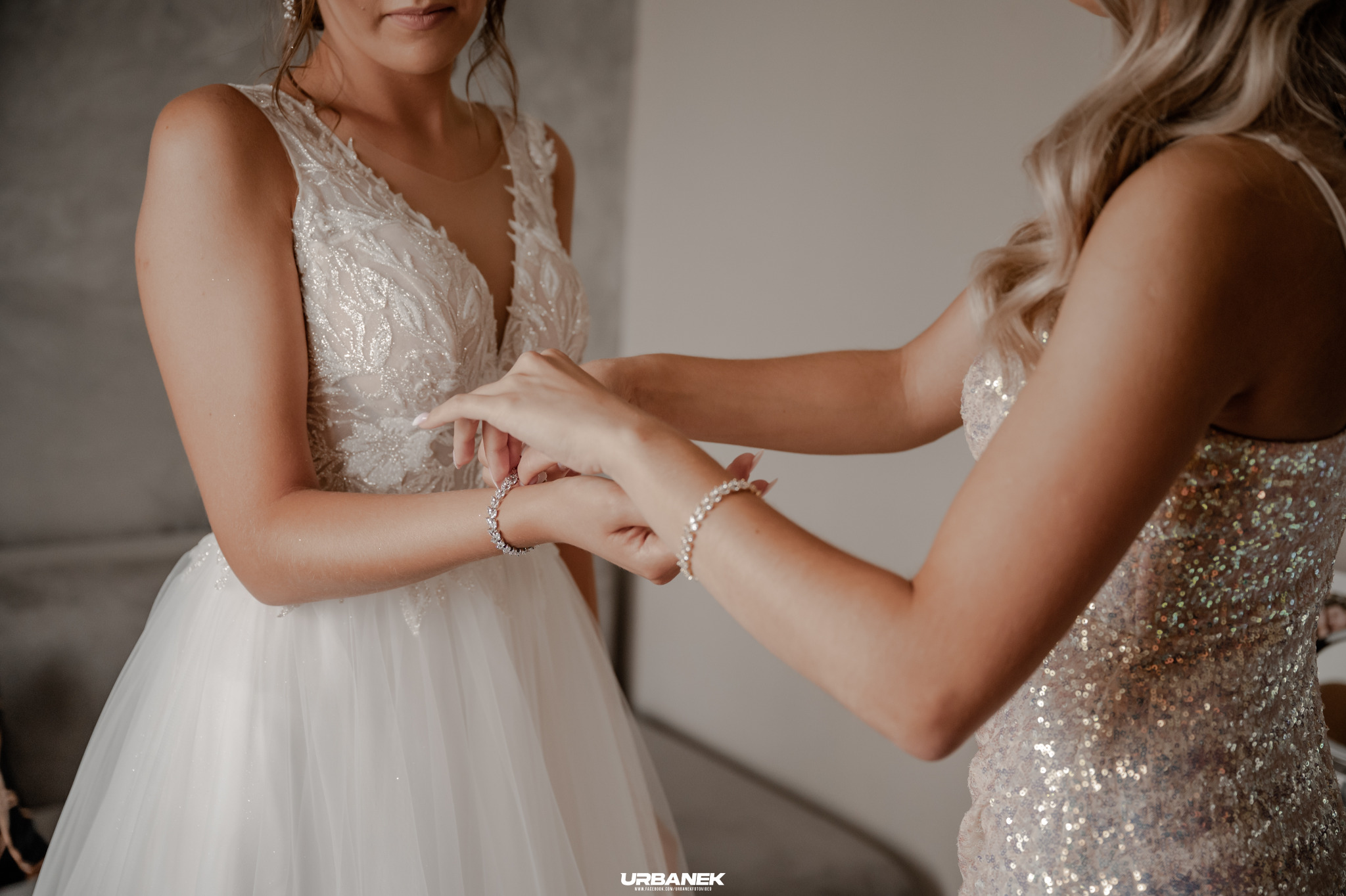 Panna młoda w białej sukni z koronkowymi aplikacjami, druhna zapina jej bransoletkę z cyrkoniami na nadgarstku.