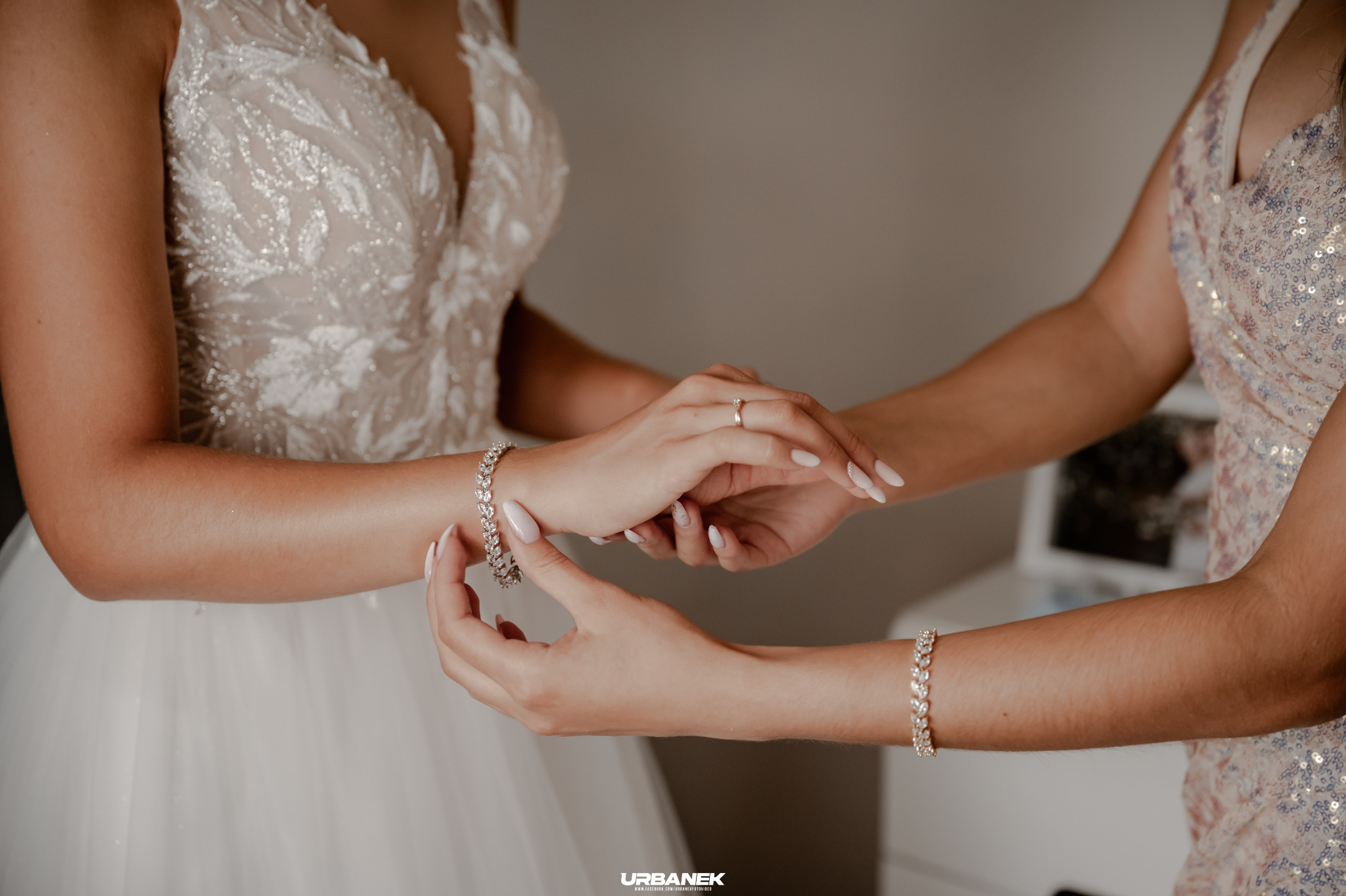 Panna młoda w białej sukni z dekoltem w serek, zakładająca błyszczącą bransoletkę, z pomocą kobiety w cekinowej sukience.