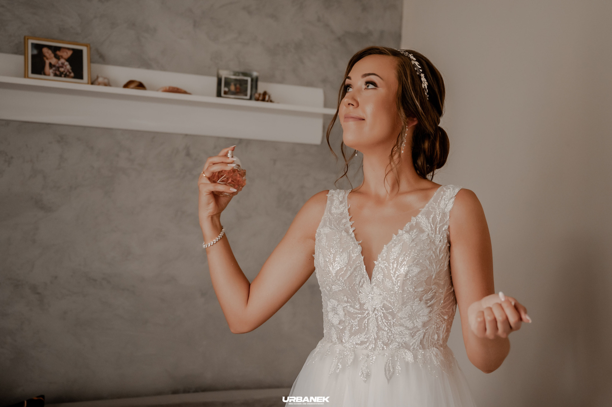 Panna młoda w białej sukni z koronkowymi zdobieniami aplikuje perfumy przed lustrem, na półce w tle widoczne pamiątki.