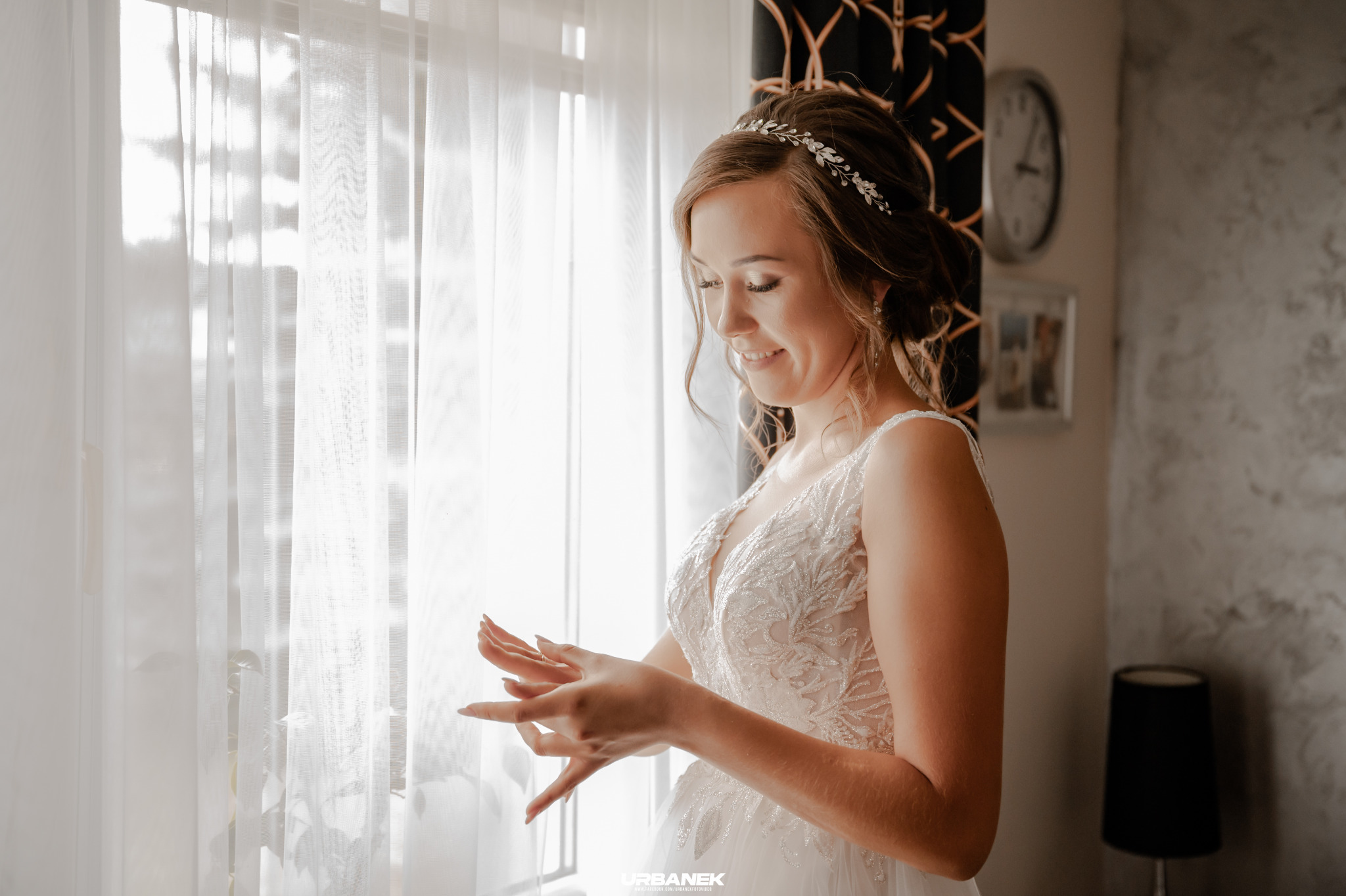 Uśmiechnięta panna młoda w białej sukni z koronkowymi zdobieniami, delikatną tiarą i kolczykami, ogląda dłonie na tle okna z białymi zasłonami.