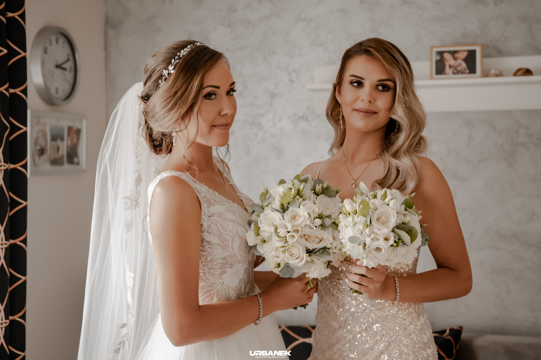Panna młoda w koronkowej sukni i welonie, trzymająca bukiet białych róż, stoi obok druhny w cekinowej sukni, obie z delikatnym makijażem i biżuterią, w tle zegar i dekoracje.
