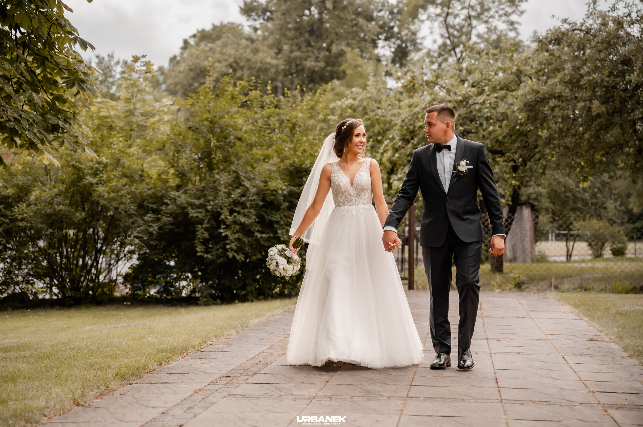 Para młoda, ona w koronkowej sukni z welonem i bukietem, on w czarnym smokingu, spacerują trzymając się za ręce po kamiennej ścieżce w parku.