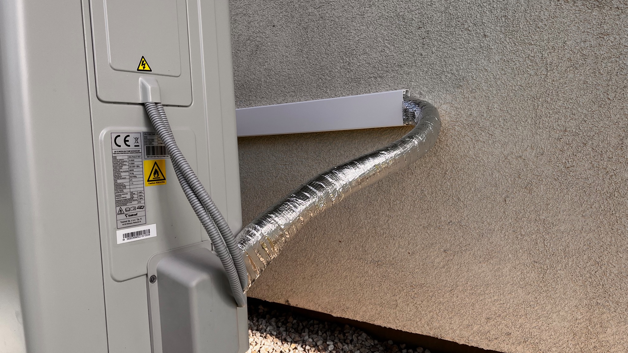Zewnętrzna jednostka pompy ciepła Galmet Prima S 10 GT zamontowana na ścianie budynku, widoczne rury w srebrnej izolacji prowadzące do wnętrza przez listwę maskującą.