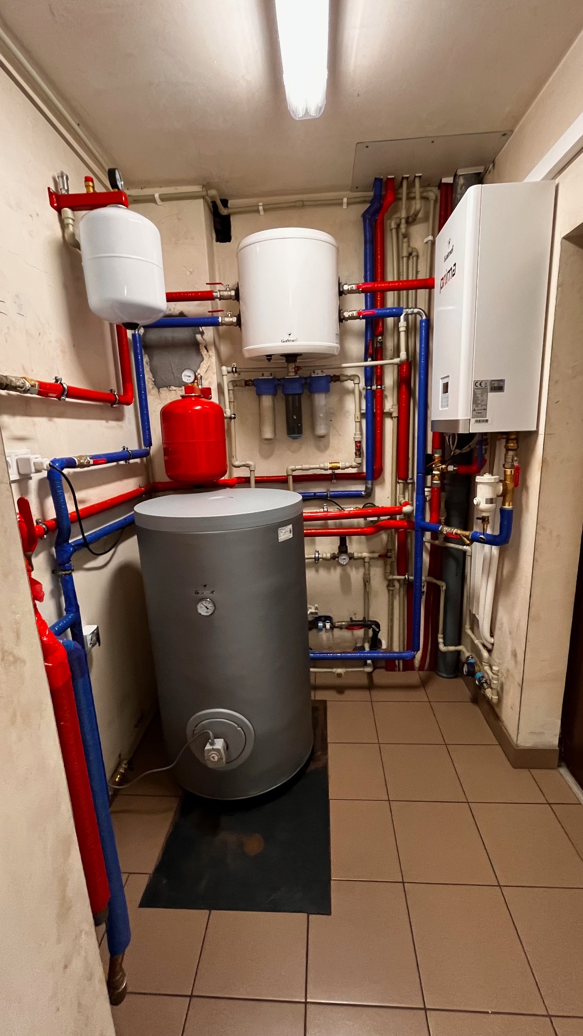 Kotłownia z szarym zasobnikiem ciepłej wody, czerwonym zbiornikiem przeponowym, białym bojlerem i wiszącym kotłem gazowym Galmet Prima S 10 GT, połączonymi rurami w kolorach czerwonym i niebieskim.