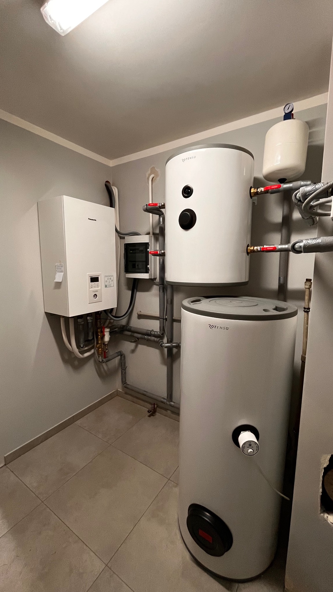 Zestaw urządzeń w kotłowni: wiszący moduł Panasonic T-Cap Split 9kW, dwa białe zbiorniki Rotenso oraz skrzynka elektryczna, połączone rurami w szarej, wykafelkowanej piwnicy.