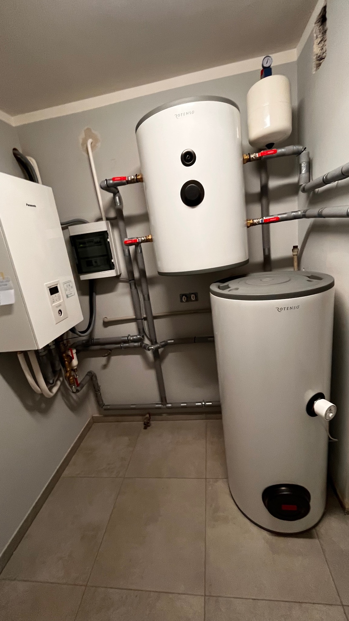 Instalacja dwóch zbiorników akumulacyjnych Rotenso, połączonych rurami, obok wiszącego, białego kotła Panasonic T-Cap Split 9kW i skrzynki elektrycznej na tle szarej ściany i płytek podłogowych.