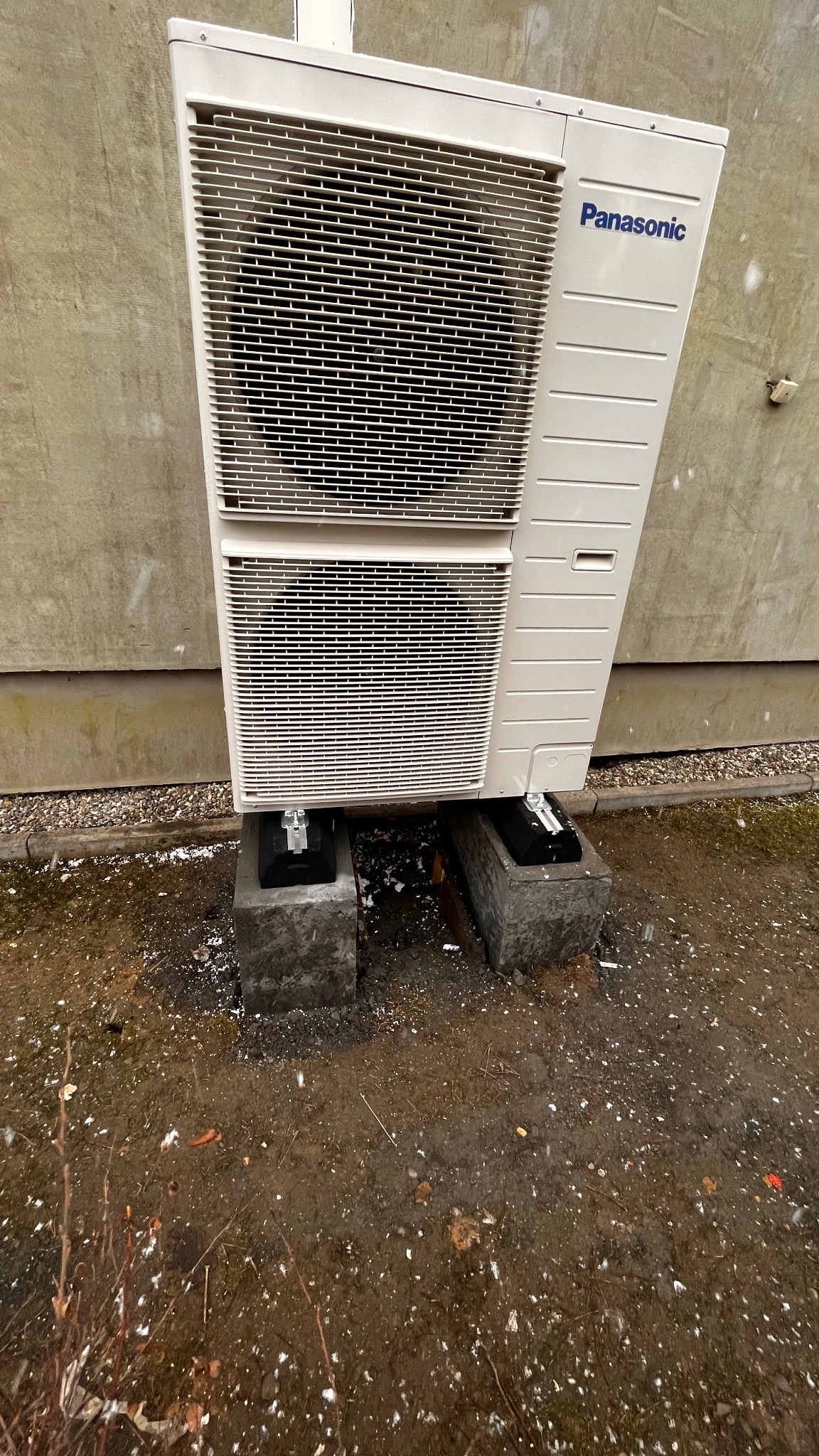 Zewnętrzna jednostka pompy ciepła Panasonic T-Cap Split 9kW, zamontowana na betonowych podstawach przy szarej ścianie budynku, widoczne opady śniegu.