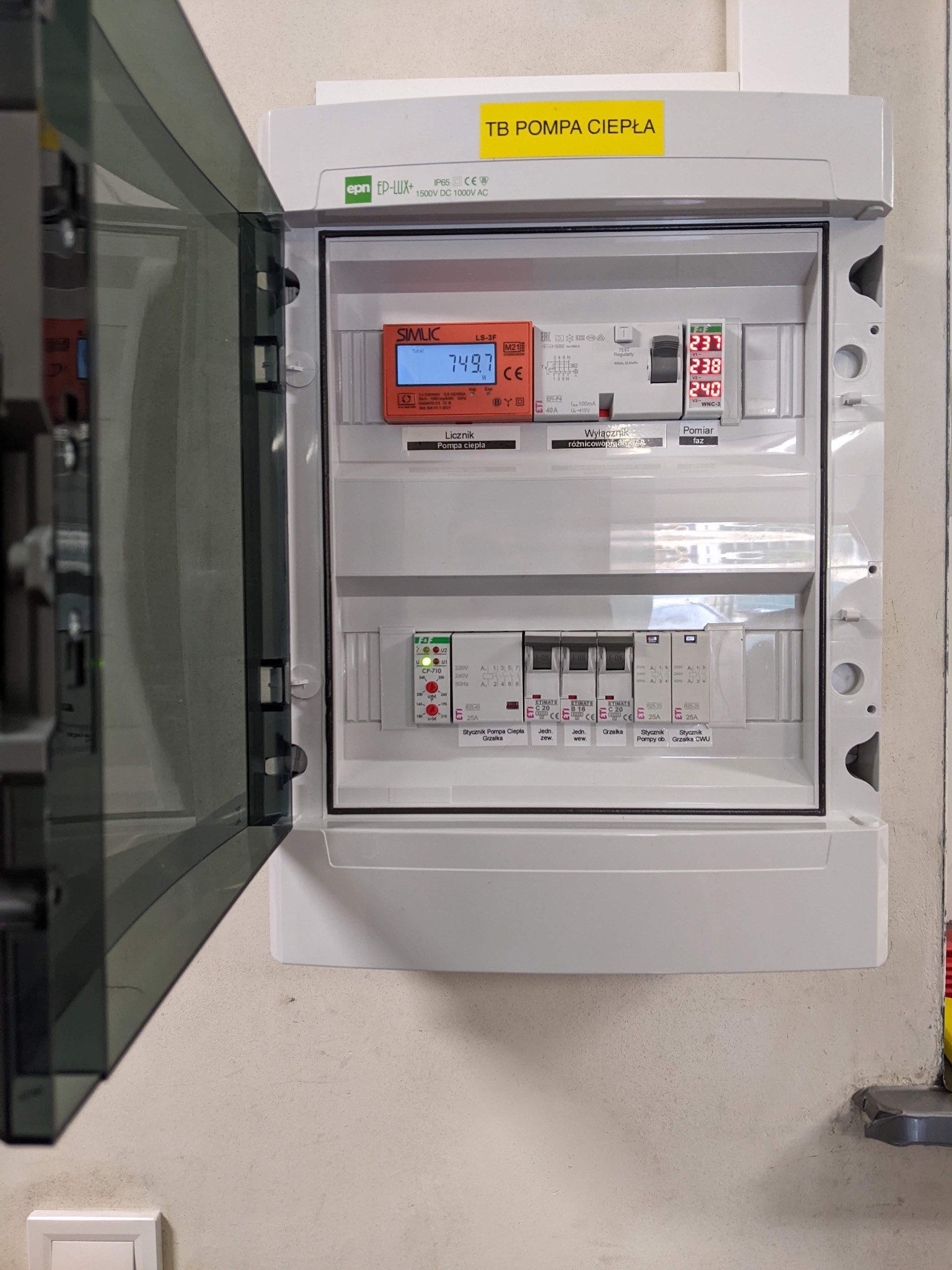 Otwarta skrzynka elektryczna z licznikiem i wyłącznikami, oznaczona żółtą naklejką 'TB POMPA CIEPŁA', widoczne wartości napięcia i zużycia energii.