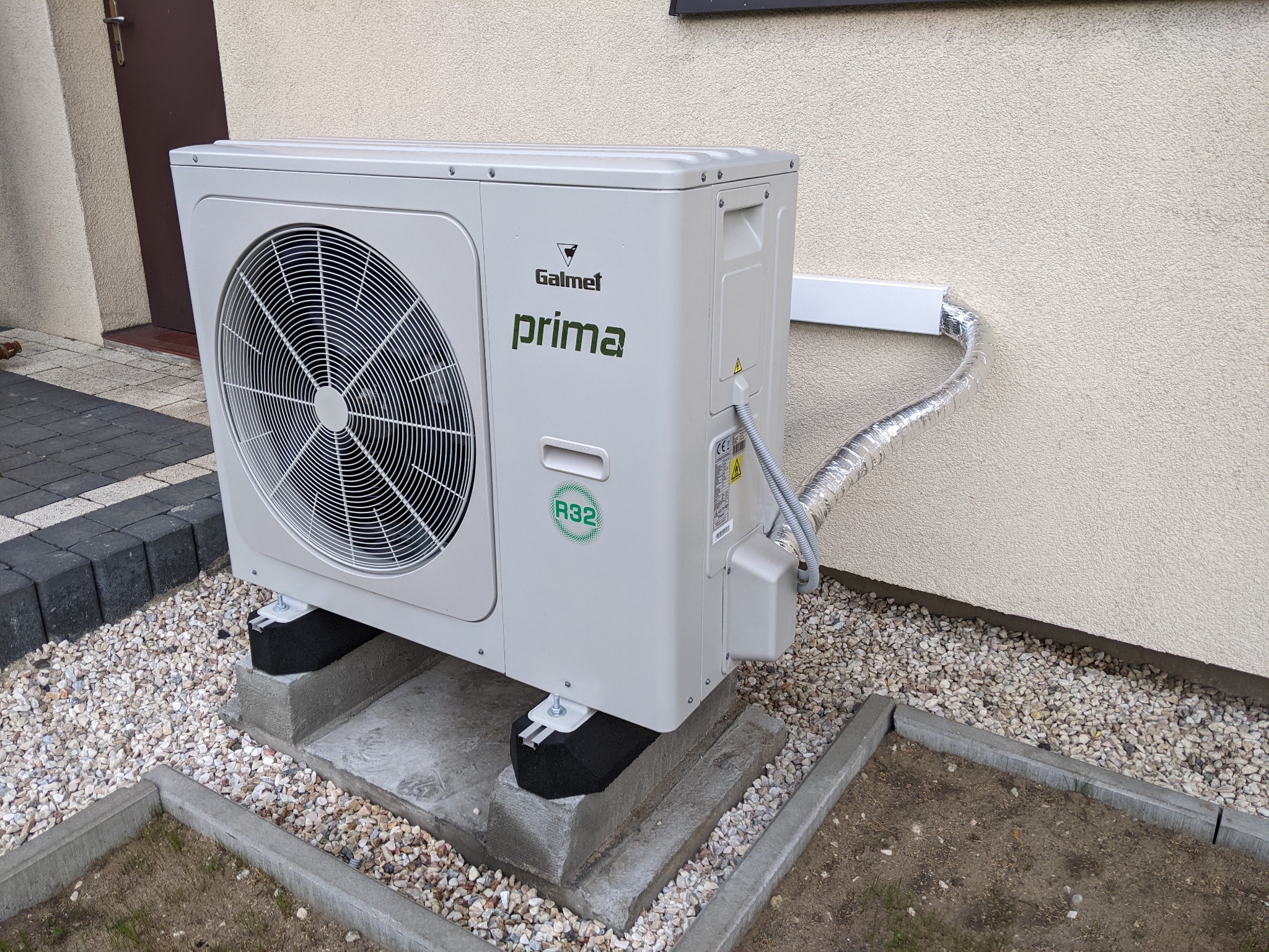 Zewnętrzna jednostka klimatyzacji Galmet Prima R32 zamontowana na betonowych bloczkach z tłumikami drgań, z widoczną aluminiową rurą odprowadzającą skropliny, na tle elewacji pokrytej jasnym tynkiem.
