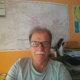 Portret mężczyzny w okularach na tle mapy miasta Wieliczka z zaznaczonymi drogami, siedzącego przy biurku z papierami i kapeluszem.