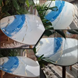 Okrągły stolik kawowy z unikalnym, ręcznie malowanym wzorem w odcieniach błękitu i złota, przypominającym marmurkowe żyłki, ustawiony na drewnianej podłodze obok wzorzystego dywanu i zielonej...