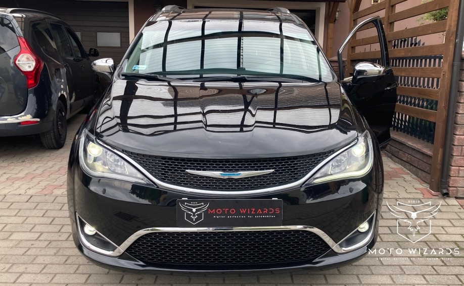 Czarny samochód Chrysler Pacifica z otwartymi drzwiami pasażera, zaparkowany na brukowanej nawierzchni przed budynkiem z drewnianym ogrodzeniem. Na przednim zderzaku widoczna tabliczka z logo 'Moto...