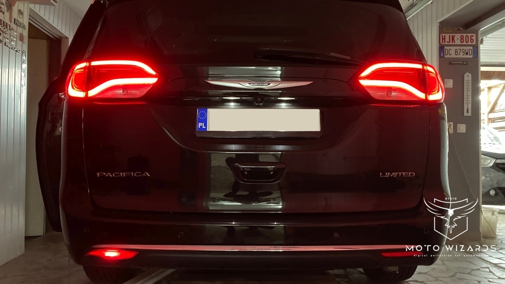 Tył czarnego samochodu Chrysler Pacifica Limited z włączonymi światłami hamowania, widoczne tablice rejestracyjne (polska i niemiecka), logo firmy Moto Wizards w prawym dolnym rogu.