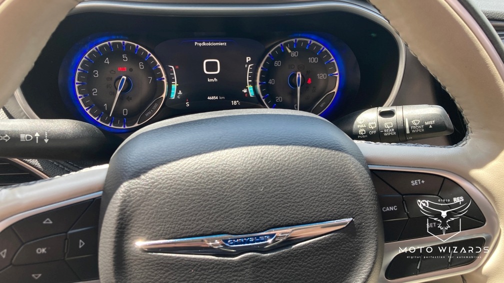 Wnętrze samochodu marki Chrysler, widok na deskę rozdzielczą z podświetlanymi zegarami, kierownicę z logo i przełączniki.