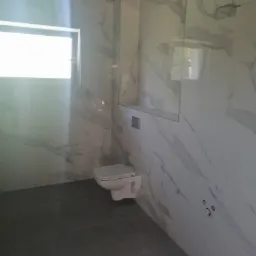 Nowoczesna łazienka w trakcie wykańczania: białe marmurowe płytki na ścianach, szara podłoga, podwieszana toaleta i okno w tle.