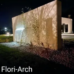 Nowoczesny ogród nocą: betonowa ściana z podświetlonym strumieniem wody, bambus i kamień ozdobny, w tle minimalistyczny budynek.