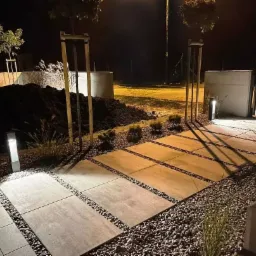 Oświetlona ścieżka z płyt betonowych i żwiru w ogrodzie nocą, widoczne młode drzewka i kopiec ziemi.