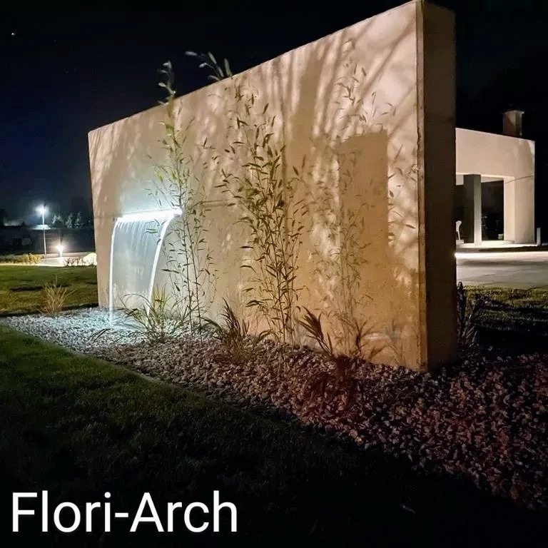 Nowoczesny ogród nocą: betonowa ściana z podświetlonym strumieniem wody, bambus i kamień ozdobny, w tle minimalistyczny budynek.