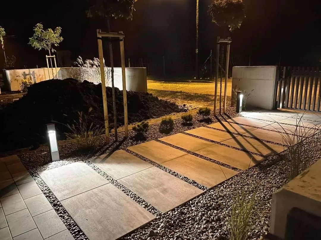Oświetlona ścieżka z płyt betonowych i żwiru w ogrodzie nocą, widoczne młode drzewka i kopiec ziemi.