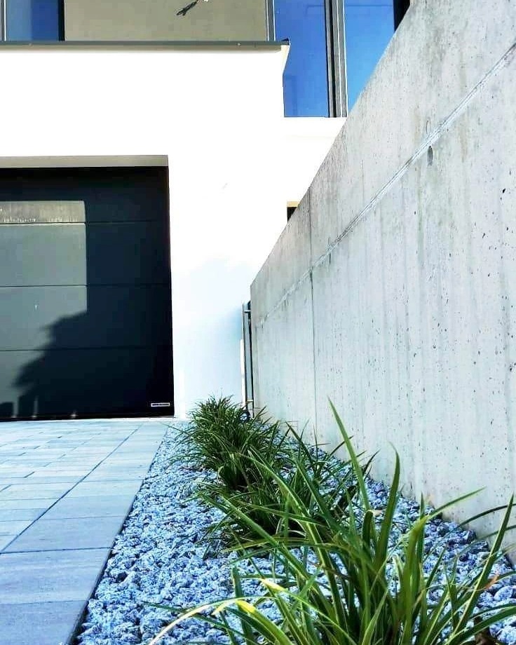 Nowoczesny ogród przy domu z szarą kostką brukową, białym budynkiem i betonowym murem, ozdobiony rabatą z niebieskich kamieni i zielonych traw.