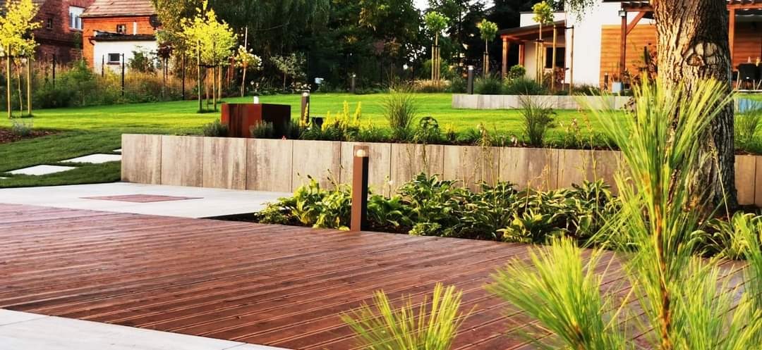 Nowoczesny ogród z drewnianym tarasem, podwyższonymi rabatami z betonowych elementów, oświetleniem punktowym i elementami architektury ogrodowej ze stali korten