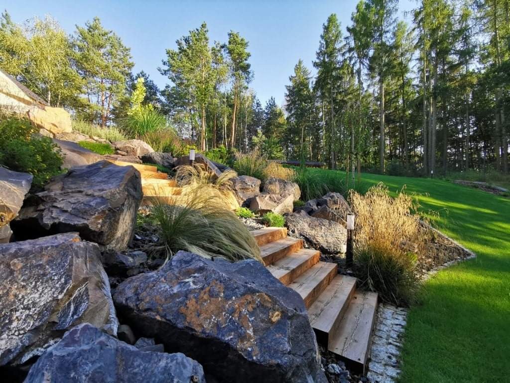 Słoneczny krajobraz ogrodu z drewnianymi schodami wkomponowanymi w skalniak z dużych, ciemnych kamieni, trawa i drzewa w tle.