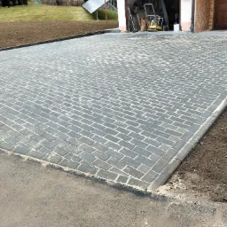 Nowo ułożona, szara kostka brukowa w jodełkę na podjeździe, z widocznym garażem i przygotowanym terenem pod trawnik. Kostka wysypana piaskiem.