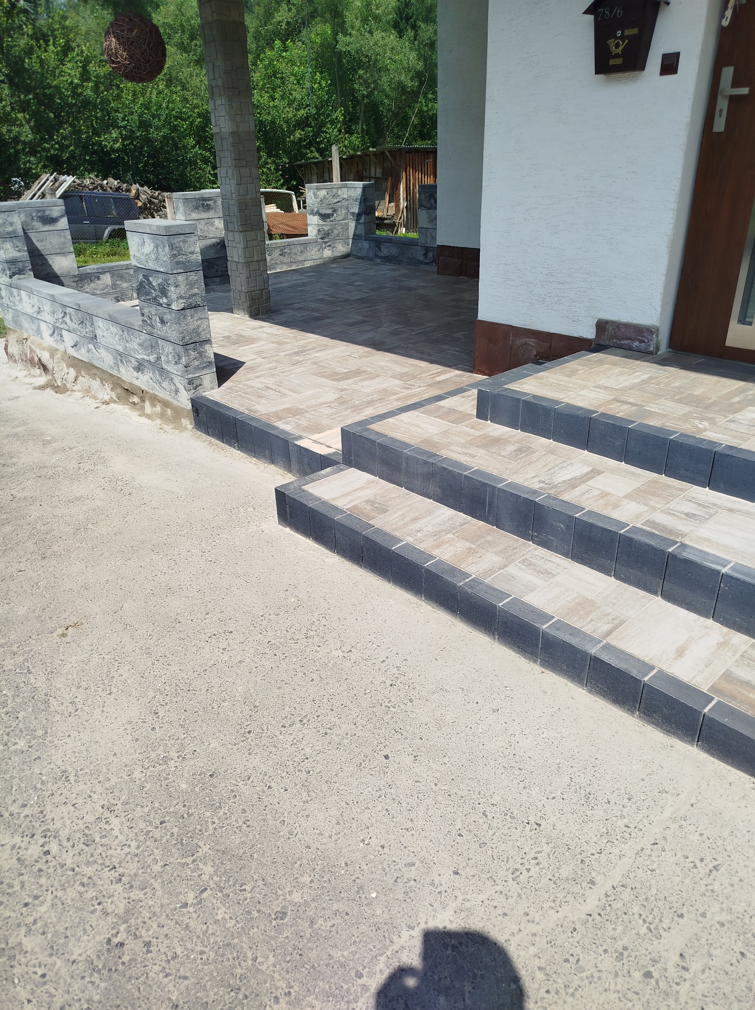 Schody wejściowe wyłożone kostką brukową w odcieniach beżu i szarości, prowadzące do drzwi wejściowych z brązową ramą. Widoczna skrzynka pocztowa i fragment podjazdu.