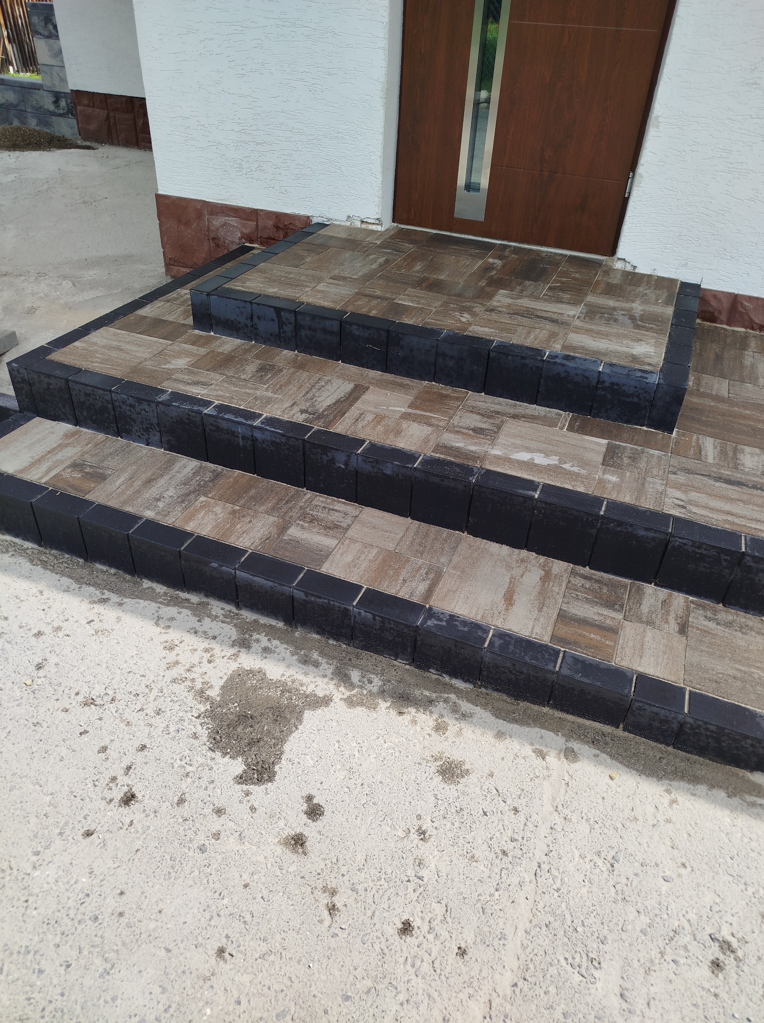 Schody wejściowe wyłożone brązowo-szarą kostką brukową z czarnym obramowaniem, prowadzące do brązowych drzwi wejściowych.