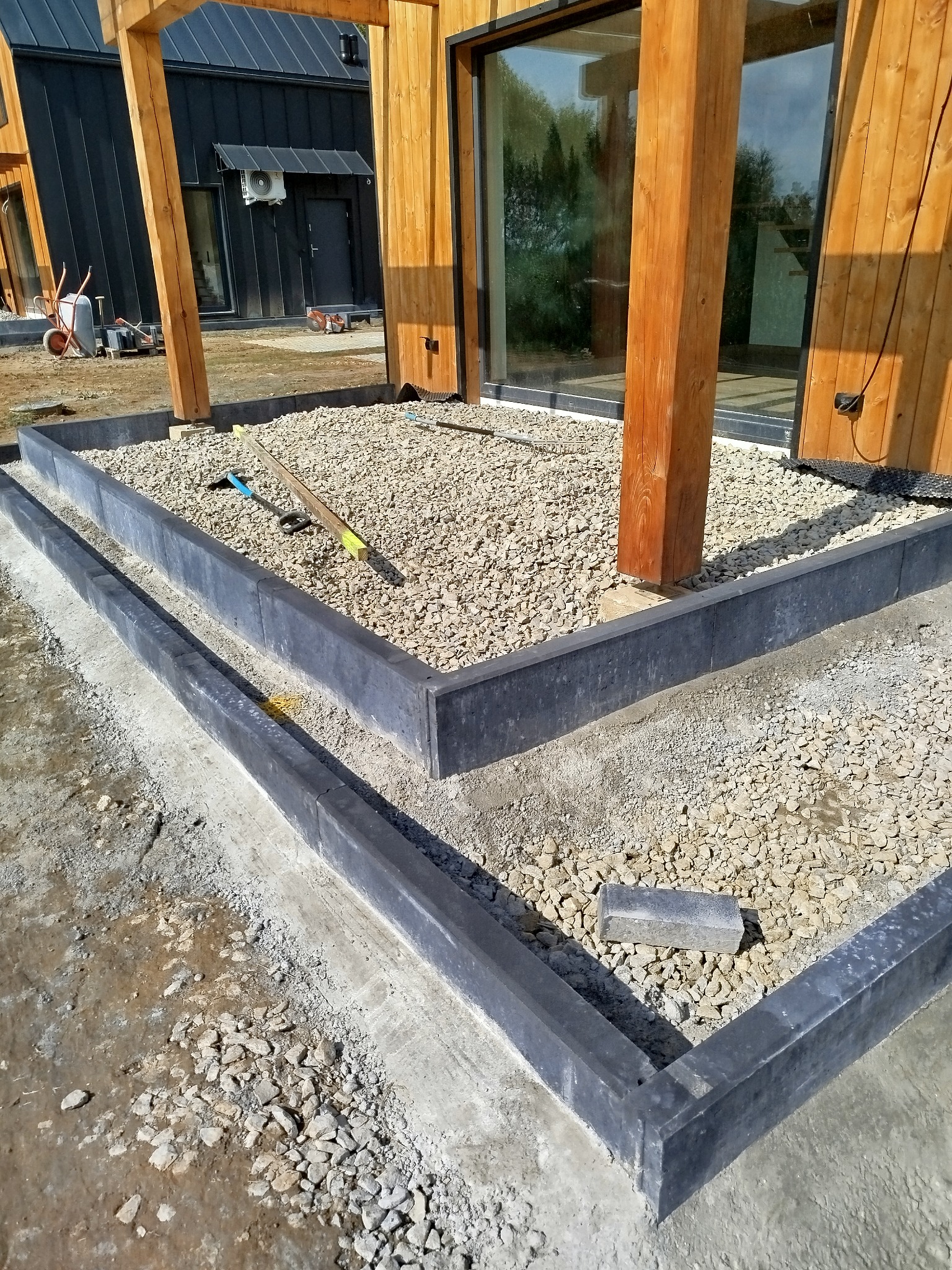 Montaż obrzeży betonowych wokół drewnianej konstrukcji domu, podsypka z drobnego kruszywa, widoczne narzędzia brukarskie.
