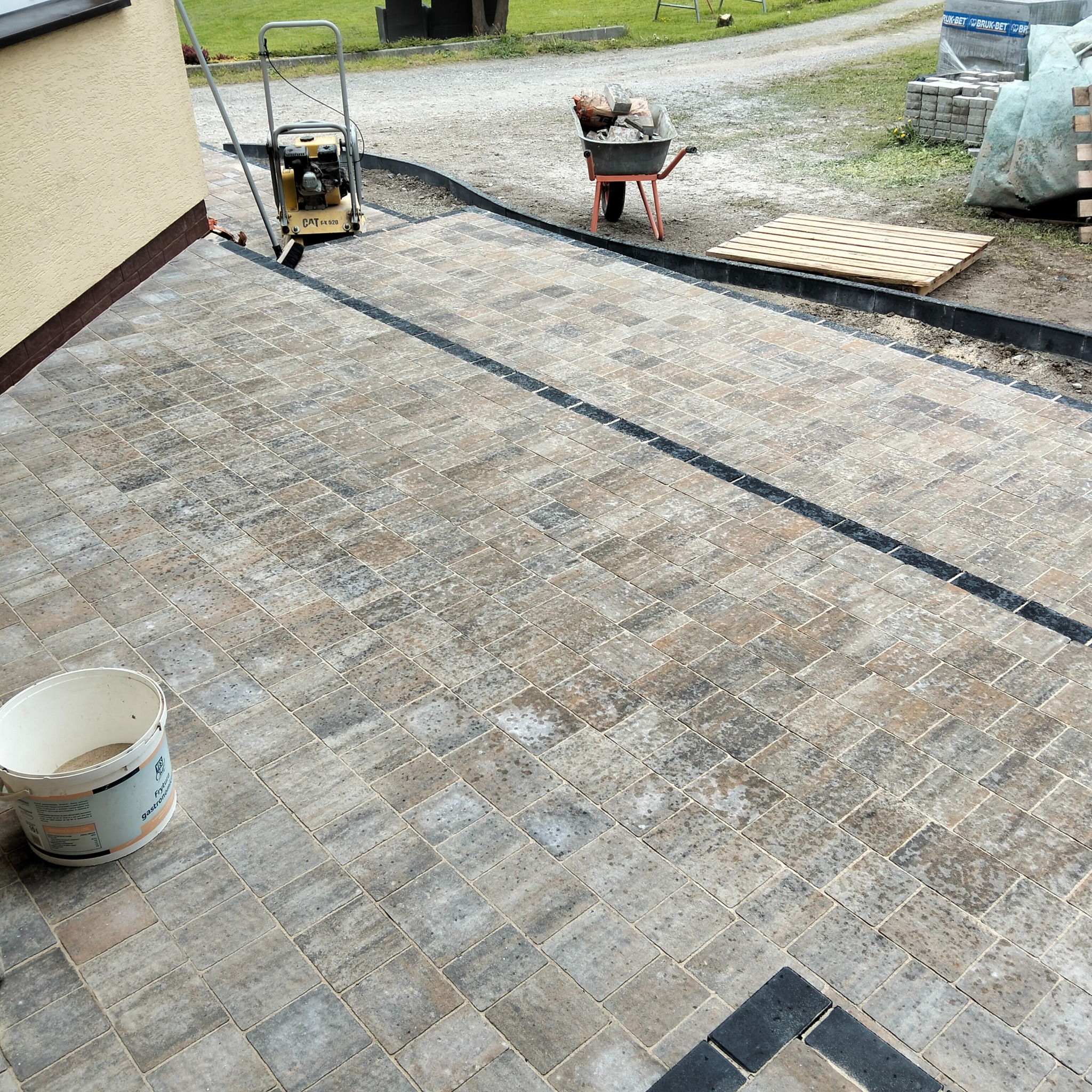 Ułożona kostka brukowa w odcieniach szarości i brązu z czarnymi akcentami. Widoczna ubijarka, taczka z materiałem i paleta. Praca w toku.